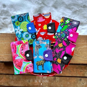 May include: Six colorful fabric card holders with different patterns, including watermelon, moose, teacups, and flowers. The card holders are arranged on a red plate with the text "Little Shop on the Tundra" printed below.