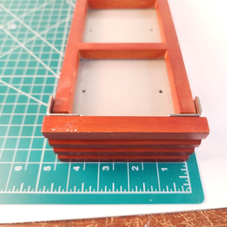 May include: A rectangular wooden frame with a reddish-brown finish, featuring two open square compartments and silver metal corner brackets. The frame is positioned on a green cutting mat with white grid lines, showing a width of 5 inches.