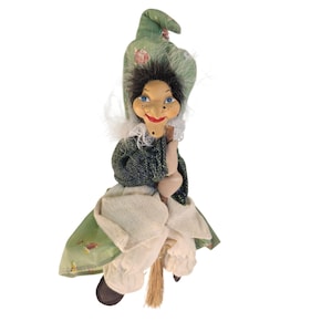 May include: A decorative witch doll featuring a green floral hat and matching coat, white trousers, and black shoes. The doll has black hair, blue eyes, and a pointed nose, and is depicted holding a small wooden broomstick with straw bristles.
