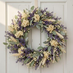 Puede incluir: Una corona circular que presenta un arreglo denso de ramas de lavanda morada, flores de hortensia color crema, hojas de oreja de cordero verde salvia y plumas de hierba de la pampa beige, creando una exhibici&oacute;n bot&aacute;nica texturizada.