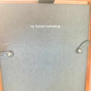 May include: The back of a square picture frame featuring a dark gray backing board with two black swivel clips and a small black hanging tab. The text Hallmark Licensing, Inc. is printed in white on the center of the backing board.