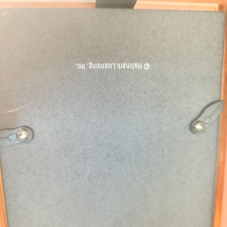 May include: The back of a square picture frame featuring a dark gray backing board with two black swivel clips and a small black hanging tab. The text Hallmark Licensing, Inc. is printed in white on the center of the backing board.