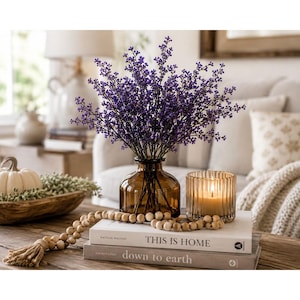 Puede incluir: Un ramo de peque&ntilde;as bayas moradas en un jarr&oacute;n de vidrio &aacute;mbar, junto a un portavelas de vidrio estriado con una vela encendida, sobre dos libros apilados titulados This Is Home y down to earth, con una guirlanda de cuentas de madera.