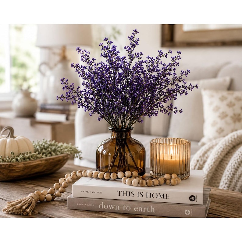 Puede incluir: Un ramo de peque&ntilde;as bayas moradas en un jarr&oacute;n de vidrio &aacute;mbar, junto a un portavelas de vidrio estriado con una vela encendida, sobre dos libros apilados titulados This Is Home y down to earth, con una guirlanda de cuentas de madera.