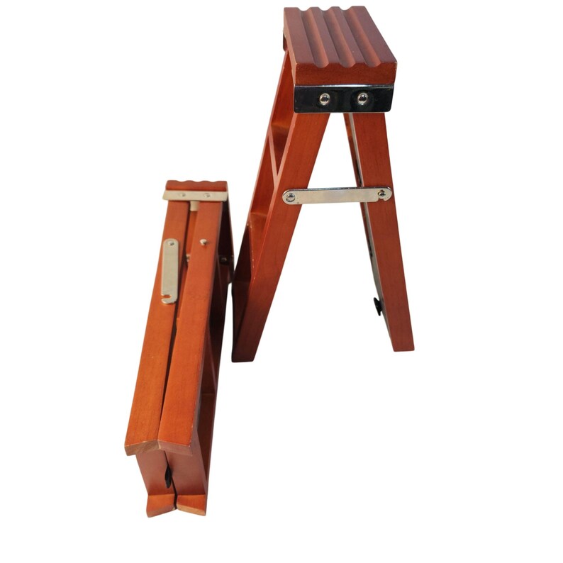 May include: Two small, reddish-brown wooden step stools. One stool is fully unfolded, showing a top platform with three grooves and silver metal hinges. The second stool is folded flat, displaying its side frame and a silver metal locking arm.
