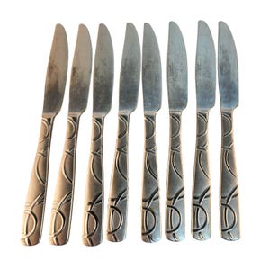 May include: Set of eight silver-colored knives with a decorative handle design. The blades are slightly curved and the handles feature an etched pattern of connected circles and lines. The knives are arranged in a row against a white background.
