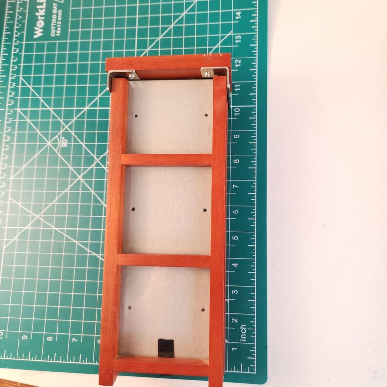 May include: A rectangular wooden frame with a reddish-brown finish, featuring three internal sections with light gray backing panels. The frame is positioned on a green cutting mat with visible inch markings, showing a length of 12 inches.