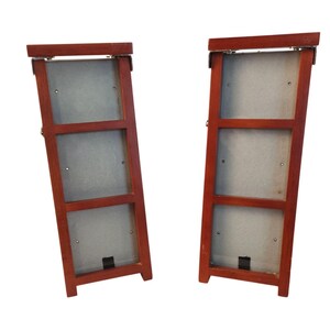 May include: Two vertical wooden picture frames with a reddish-brown finish. Each frame features three rectangular openings arranged in a column, with light gray backing boards visible through the openings and metal hinges at the top edges.