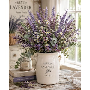 Puede incluir: Una vasija de cer&aacute;mica color crema con dos asas laterales, que presenta el texto "FRENCH LAVENDER FARM" y un gr&aacute;fico floral. Est&aacute; llena de un arreglo grande y denso de ramas de lavanda morada, peque&ntilde;as margaritas blancas y hojas de eucalipto.