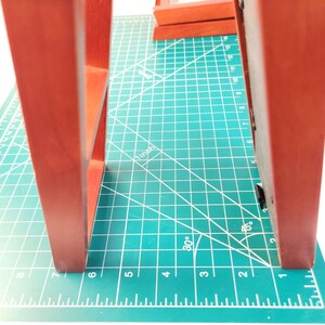 May include: Two reddish-brown wooden picture frames standing upright on a green cutting mat. The frames feature a smooth finish and a rectangular structure. A visible 1-inch measurement mark is printed on the mat between the two frames.