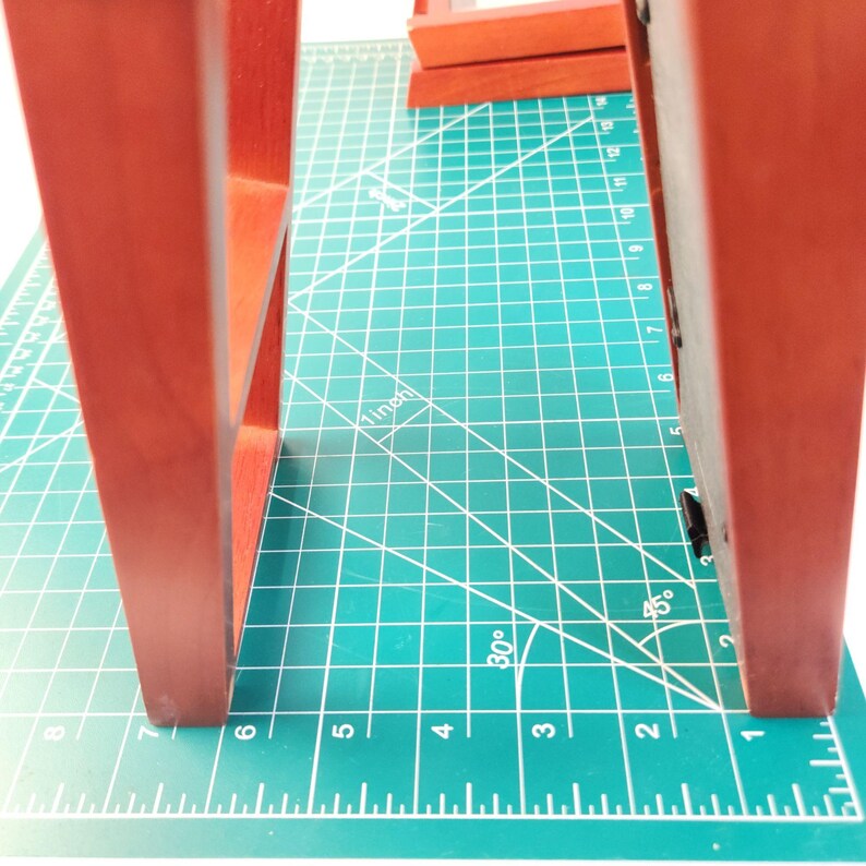 May include: Two reddish-brown wooden picture frames standing upright on a green cutting mat. The frames feature a smooth finish and a rectangular structure. A visible 1-inch measurement mark is printed on the mat between the two frames.