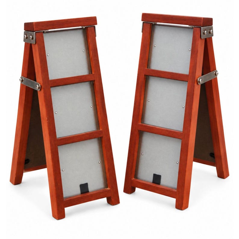 May include: Two A-frame picture frames constructed from reddish-brown wood, each featuring three vertical rectangular openings with silver-toned metal backing plates. The frames are connected by silver-colored metal hinges at the top and sides.