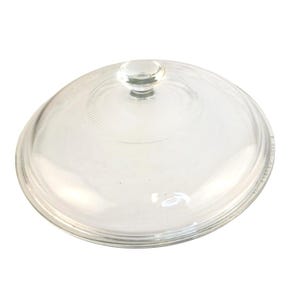 May include: A clear glass lid with a round knob handle. The lid is slightly domed and has a circular pattern etched into the glass. The lid is designed to fit on a pot or pan.