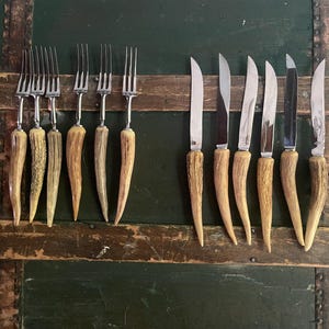 May include: A set of eight vintage knives and forks with antler handles. The knives and forks have silver blades and are arranged in two rows on a wooden surface.