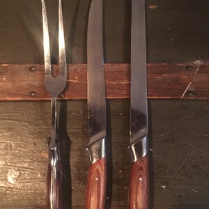May include: Three kitchen utensils with wooden handles: a fork and two knives. The utensils are silver with a polished finish. The wooden handles are a dark brown color.