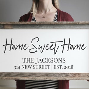 Pode incluir: Uma placa de madeira branca com o texto "Home Sweet Home" em escrita cursiva preta. Abaixo do texto est&aacute; "THE JACKSONS", "314 NEW STREET" e "EST. 2018".