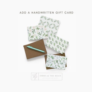 May include: A set of blank greeting cards with a floral design. The cards are white with green and white floral patterns. The cards are stacked on a brown surface. The text "ADD A HANDWRITTEN GIFT CARD" is at the top of the image. The text "EMMA & THE BEAN personalized gifts" is at the bottom of the image.
