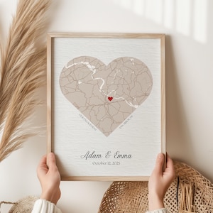 May include: A framed print featuring a heart-shaped map in neutral tones, with a red heart marking a location. The print includes the names "Adam & Emma" and the date "October 12, 2025". The frame is light-colored wood.