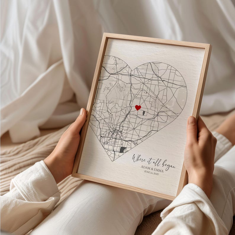 May include: A framed print with a heart-shaped map of a city. The map is in black and white with a red heart marking a specific location. The text below the map reads "Where it all began, Adam & Emma, June 14, 2023".