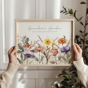May include: A framed print featuring a watercolor illustration of a garden with various flowers in shades of purple, yellow, orange, and pink. The text "Grandma's Garden" is written at the top, with names below. The frame is made of light-colored wood.