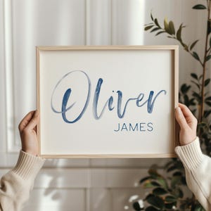 May include: A framed print featuring the name "Oliver" in a flowing, dark blue script above the name "JAMES" in a smaller, sans-serif font. The artwork is set against a cream-colored background within a light wood frame.