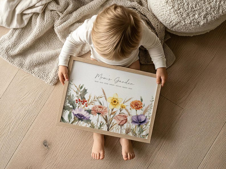 May include: A framed print with the text "Mom's Garden" and floral illustrations in shades of purple, pink, yellow, and orange. The print is held by a person on a light wood floor, with a neutral-toned blanket and pillow in the background.