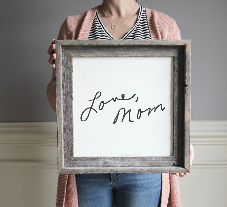 May include: A framed sign with a white background and black handwritten text that reads "Love, Mom". The frame is made of weathered wood.