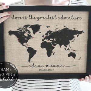 May include: A black framed burlap map of the world with the text "Love is the greatest adventure" at the top. The map has red push pins marking locations. The text "adam emma 10.29.2017" is at the bottom of the map.