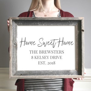 Pode incluir: Uma placa enquadrada com o texto "Home Sweet Home" em escrita cursiva. Abaixo do texto est&aacute; "The Brewsters", "8 Kelsey Drive", e "Est. 2018". A placa &eacute; segurada por uma pessoa em frente a uma parede cinza.