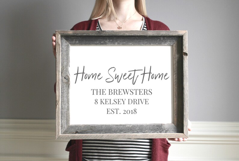 Pode incluir: Uma placa enquadrada com o texto "Home Sweet Home" em escrita cursiva. Abaixo do texto est&aacute; "The Brewsters", "8 Kelsey Drive", e "Est. 2018". A placa &eacute; segurada por uma pessoa em frente a uma parede cinza.