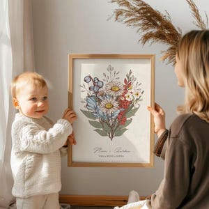 May include: A framed floral print featuring a bouquet of daisies, blue flowers, and red accents. The artwork is in a wooden frame and includes the text "Mum's Garden" and names. The print is held by a person and a child.