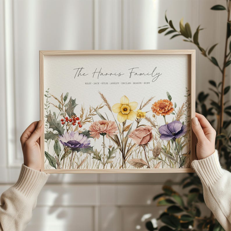 May include: A framed floral print featuring a watercolor illustration of various flowers in shades of purple, yellow, orange, and pink. The text "The Harris Family" is displayed above the flowers, with family names below. The frame is made of light-colored wood.