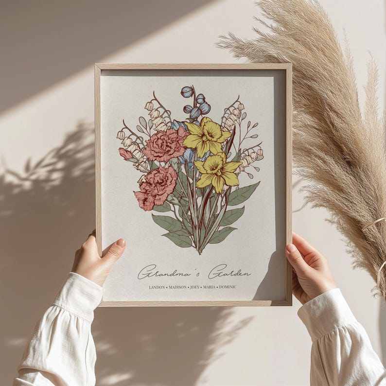 May include: A framed floral print featuring a bouquet of pink carnations, yellow daffodils, and blue accents. The print is labeled "Grandma's Garden" with names below. The artwork is held by hands and has a light wood frame.