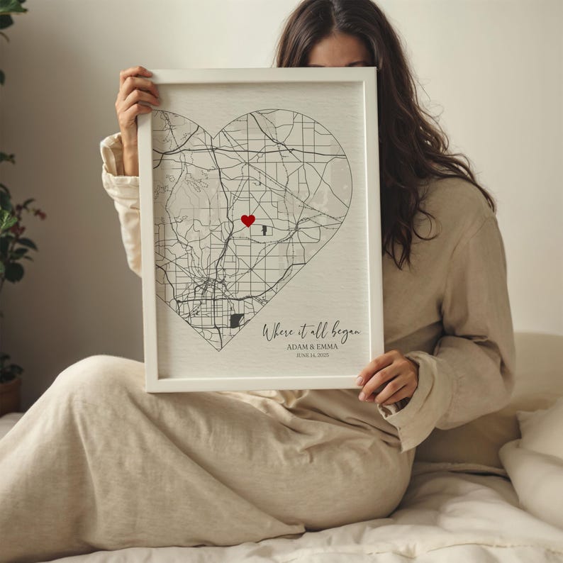 May include: A white framed print with a black and white map of a city in the shape of a heart. A red heart is placed on the map. The text "Where it all began" is written below the map with the names "Adam & Emma" and the date "June 24, 2020".