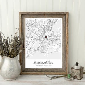 May include: A framed black and white map of New York City with a red house icon marking a location. The text below the map reads "Home Sweet Home" with the names "Adam & Emma" and the date "EST. 2022".