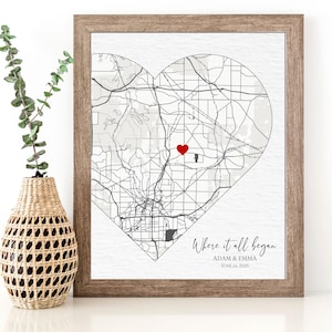 May include: A framed print with a heart-shaped map in black and white with a red heart marking a location. The text below the map reads "Where it all began, Adam & Emma, June 14, 2023".