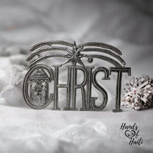 May include: Silver metal ornament with the word "CHRIST" in large letters, with a star and a depiction of the nativity scene above the word. The ornament is on a white fabric background.