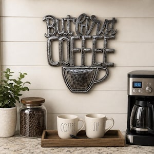 May include: A dark metallic wall sign featuring the text But first COFFEE with a coffee cup graphic. The letters and cup have a hammered texture and a beaded border. The sign is mounted on a white wall above a wooden tray with two mugs.