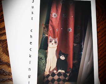 May include: A greeting card with the text "just checking!" features a whimsical illustration of three cats. One cat peeks from behind a red curtain, while two others sit on a checkered cushion. The card is on a wooden surface.