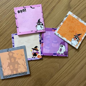 May include: A collection of square Halloween-themed sticky notes. Each note features a different design, including ghosts, witches, bats, and haunted houses. The color palette includes pink, orange, purple, and gray, with the word "BOO!" on one note.
