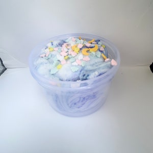 May include: A clear plastic container filled with light blue, white, and purple slime with yellow and pink star and crescent moon shaped sprinkles.