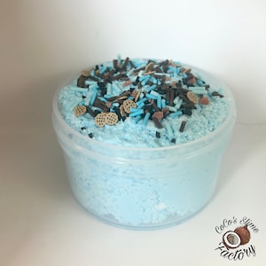 May include: A clear plastic pot filled with light blue slime, decorated with blue, brown, and tan sprinkles and cookie-shaped embellishments. The pot is labelled "Coco's Slime Factory".