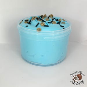 May include: A clear plastic container filled with light blue slime. The slime is topped with small, brown, and black sprinkles, and small cookie-shaped decorations. The Coco's Slime Factory logo is visible in the bottom right corner.