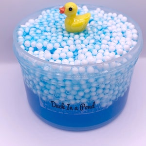 May include: Blue slime with white foam balls and a yellow rubber ducky on top. The slime is in a clear plastic container with the label "Duck In a Pond".
