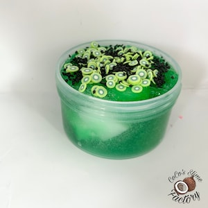 May include: A clear plastic container filled with green slime topped with black sprinkles and green kiwi slices. The container has a white lid. The slime is a light green color and has a sparkly texture. The kiwi slices are green and have a black center. The sprinkles are black and are scattered randomly on top of the slime.
