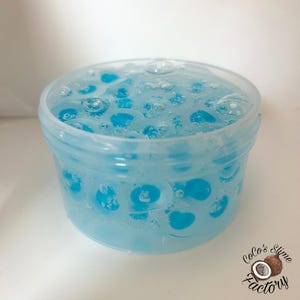 May include: A clear plastic container filled with blue slime. The slime has a bubbly texture with blue spheres suspended within. The container is labeled "Coco's Slime Factory" in a cursive font with a coconut graphic.