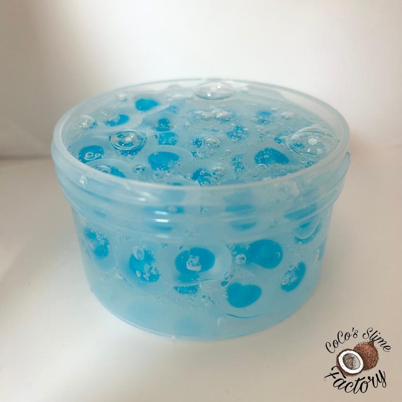 May include: A clear plastic container filled with blue slime. The slime has a bubbly texture with blue spheres suspended within. The container is labeled "Coco's Slime Factory" in a cursive font with a coconut graphic.