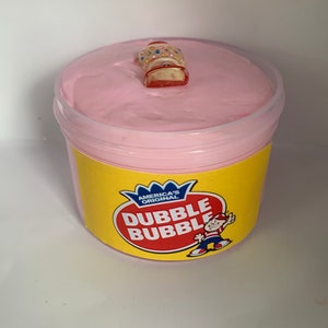 May include: Pink slime in a clear container with a yellow label that says "America's Original Dubble Bubble". A small gumball machine charm is on top of the slime.