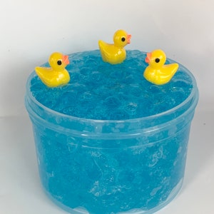 May include: A clear plastic container filled with blue slime with three yellow rubber duckies floating on top.