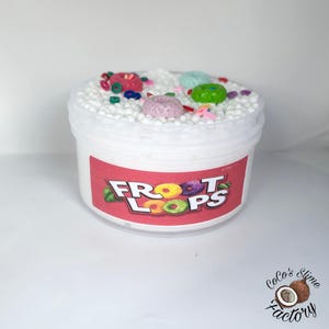 May include: A white plastic container of slime with a red label featuring the text "Froot Loops" and colorful cereal graphics. The slime is topped with white foam balls and colorful, miniature cereal pieces.